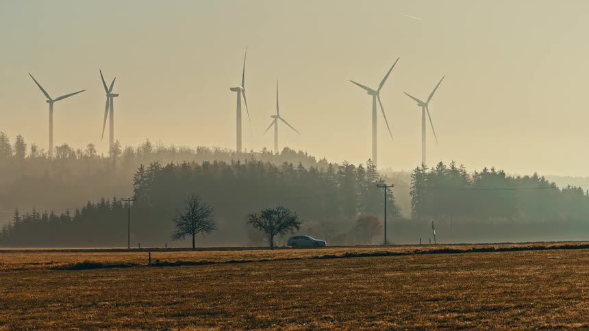 A car drives through the countryside passing open fields with a series of wind turbines spinning in the background