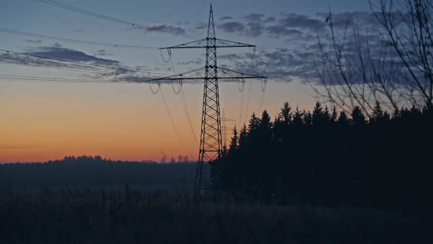A metal power tower stands in a field next to a dark forest under a blue and orange sky at dawn