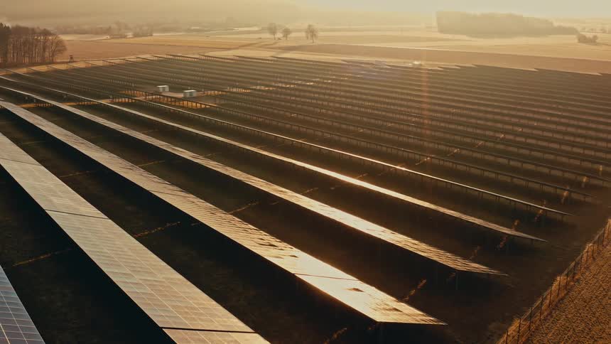 An aerial camera pans over rows of solar panels in a large field during a hazy sunrise capturing the orange light reflecting off the glass surfaces while misty hills and sparse trees occupy the background of the rural landscape