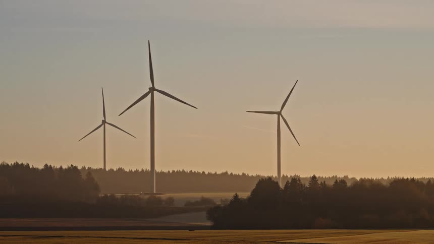 Powerful wind turbines stand over a misty forest and open field at golden sunrise