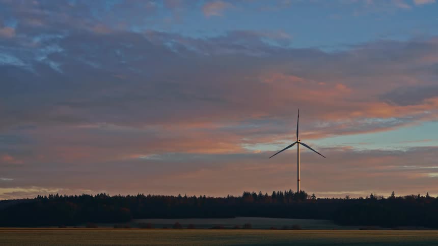 Wind turbine spins high above a forest under a cloudy sky at dusk