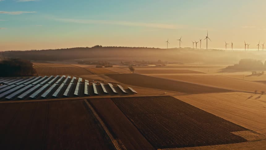 Rural farmland with solar panels lined up across the ground and wind turbines spinning on the horizon