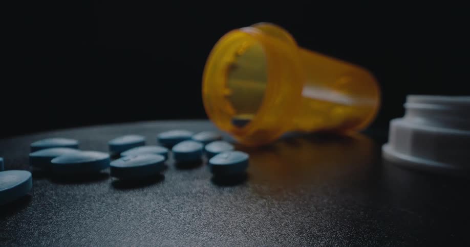 A close view pans across blue oval pills scattered on a black surface next to an open orange prescription bottle lying on its side near a white cap