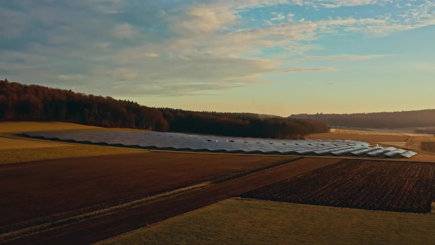 An aerial view flies over rows of solar panels situated in a brown field near a forest edge under a cloudy sky during sunset