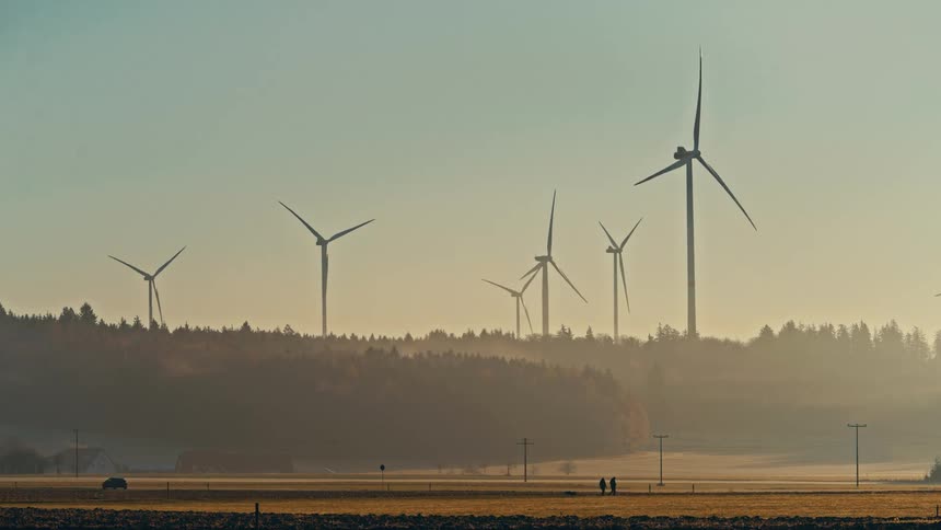 Wind turbines installed in a rural area with open fields and forest in the background under soft light