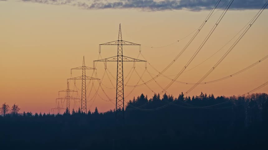 Electric power transmission lines stretching over a silhouetted forest beneath an orange dusk sky