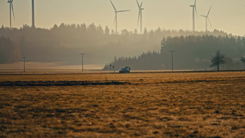 Wind turbines tower over a conifer forest on a hazy day while a person walks around to open the back of a vehicle