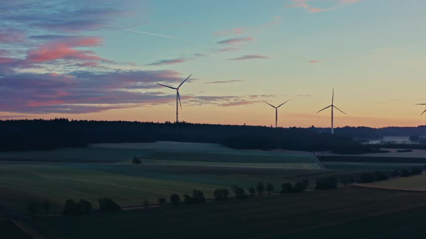 A slow aerial shot pans across a serene rural landscape where silhouetted wind turbines spin against a colorful sky at dawn