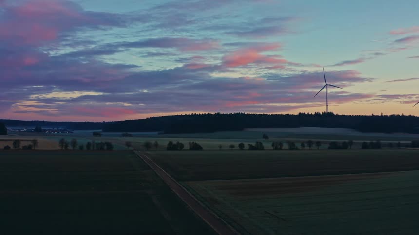An aerial drone shot pans across a rural landscape at dawn showing rotating wind turbines on a wooded hill overlooking damp agricultural fields under a streaked pink and blue sky