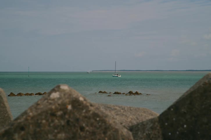 A distant white sailboat floats on calm turquoise water beyond a foreground of blurred concrete rocks under a cloudy sky