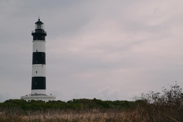 A tall black and white striped lighthouse stands prominently against a cloudy sky surrounded by low green shrubbery and dry coastal grasses on a flat horizon