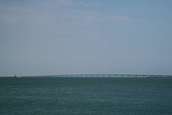 A distant wideangle view shows a long lowprofile bridge stretching across a calm body of water toward a small lighthouse under a clear blue sky