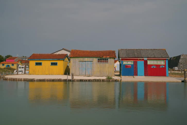 Three vibrant wooden huts in yellow tan and blue stand along a calm waterfront their colorful facades and tiled roofs reflecting clearly on the surface of the water under a soft overcast sky