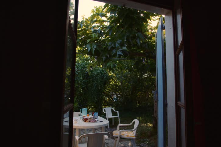 The camera looks through an open doorway onto a quiet backyard patio where a white table with drinks and snacks sits surrounded by plastic chairs under the thick green canopy of a large tree with a soft sunlight