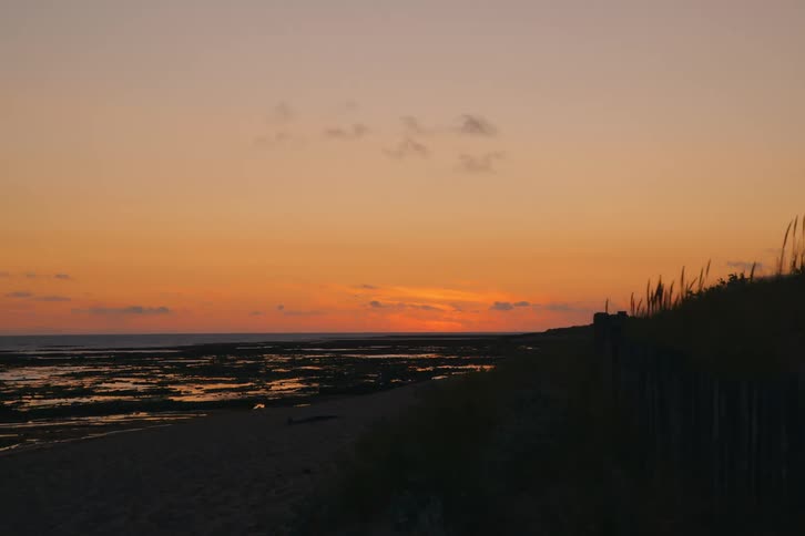 Sun sets over calm sea as wooden fence lines grassy dune
