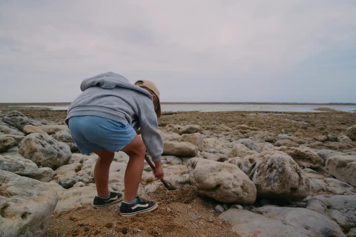 A child wearing a grey hoodie and a tan cap uses a stick to dig in a small patch of sand among large rocks on a coastline under an overcast sky