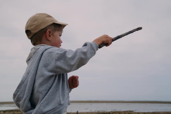A young boy in a gray hoodie and tan cap stands in a coastal wetland holding a long stick like a wand pointing it toward the horizon against a cloudy sky while looking out over the shallow water and muddy terrain