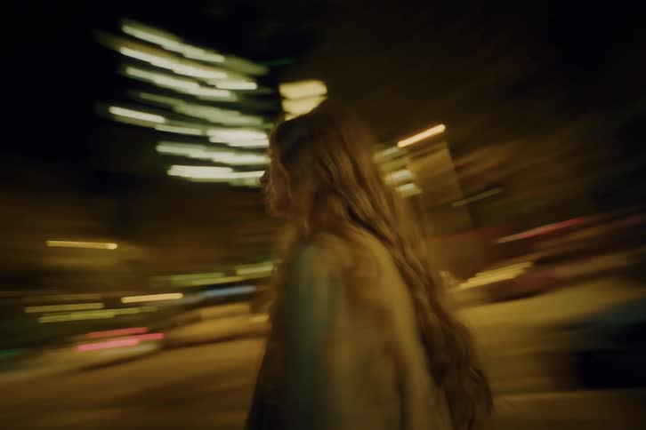 A young woman with long wavy hair spins around rapidly in the middle of a city street at night while blurred lights from buildings and traffic streak across the background