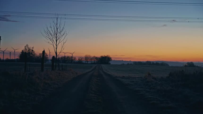 Dirt road between fields and wind turbines under orange sky with scattered trees