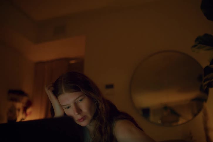 Tired woman rests their head on one hand while looking at a screen in a lowlit indoor setting