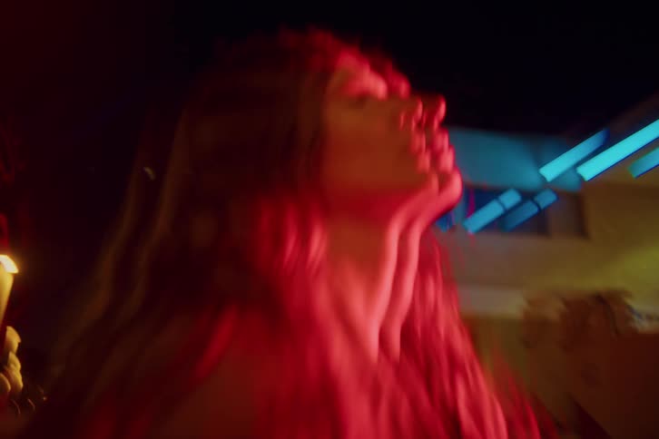A blurry view of a woman with long hair illuminated by red neon lights at night as she moves through a city with glowing building lights in the background