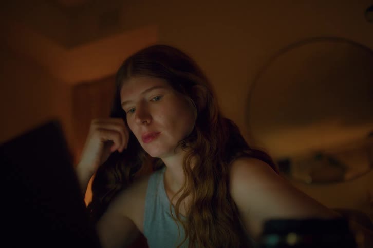 A young woman with long hair sits at a desk in a dimly lit room looking tired as she stares at her laptop screen and rests her head on her hand