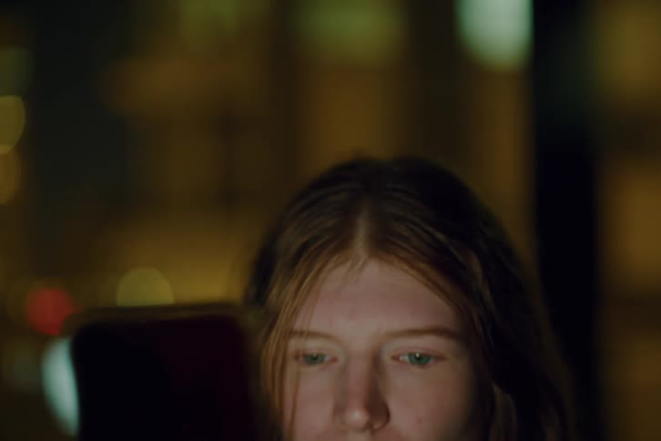 A young girl with long hair looks down at her smartphone while scrolling through content at night her face illuminated by the screens glow against a blurry background