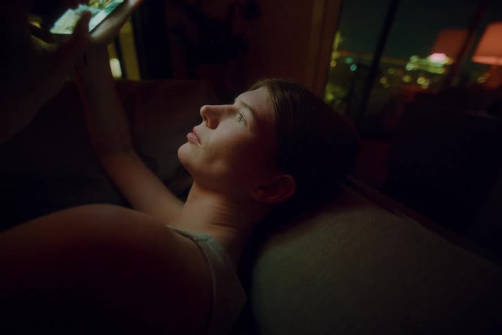 Woman lying on the sofa and scrolling on a smartphone at night at her urban apartment.