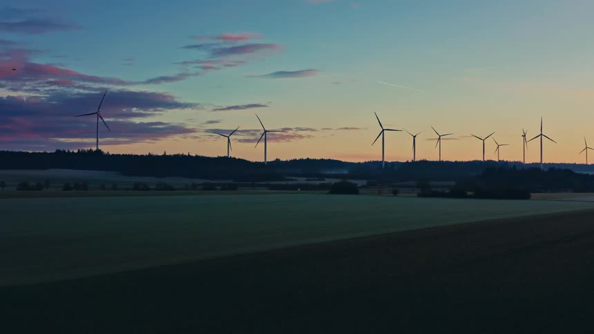 A line of industrial wind turbines stands across a ridge during a colorful sunset as the camera pans slowly over green agricultural fields and distant dark forests under a sky filled with blue and pink clouds