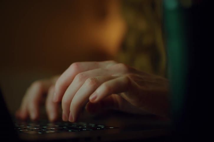 A person types on a computer keyboard in a dark room the faint glow of the screen illuminating their hands as they work in the quiet shadows