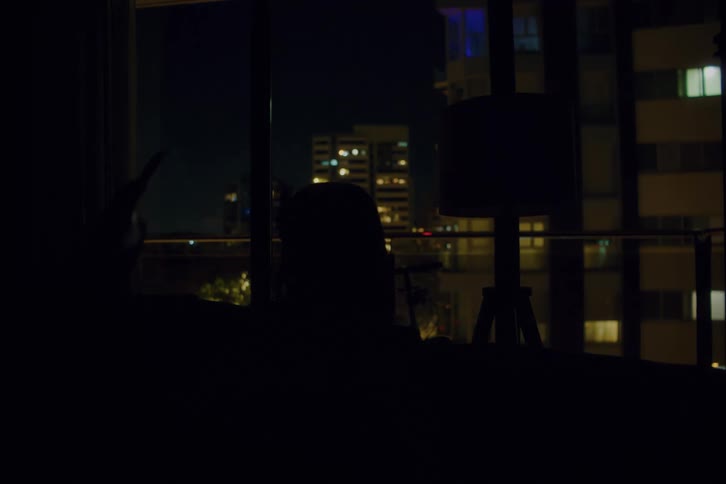 A woman with red hair sits on a sofa in a dark apartment at night taking a selfie with her smartphone while city buildings are visible through the window behind her