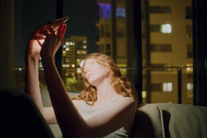 Close up of a young woman taking phone selfies while sitting on a couch indoors at night