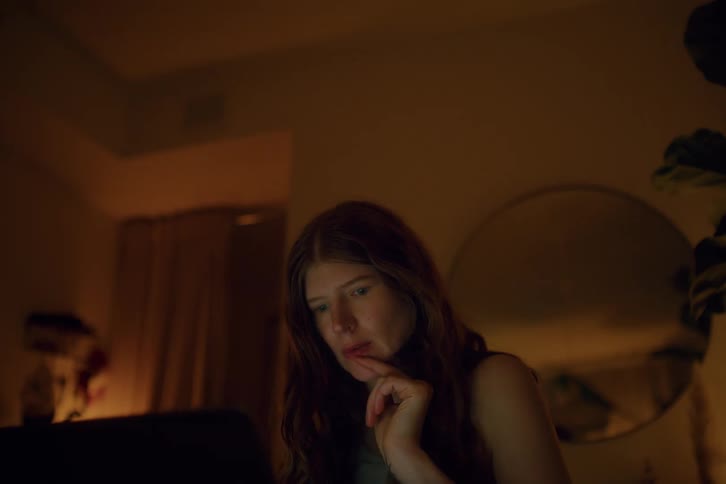 A close view of a woman with long hair focused looking at a laptop screen in a dimly lit room at night as she rests her chin on her hand