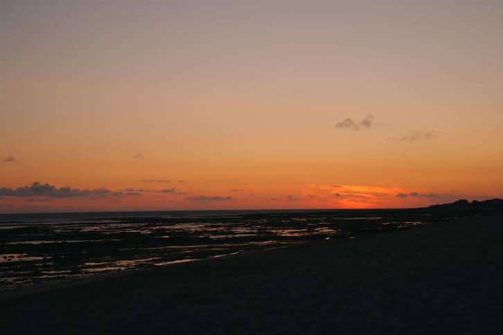 A wide static view of a rocky beach at dusk as the sun dips below the horizon casting a deep orange glow across the sky and reflecting on the shallow tide pools and dark coastal sand