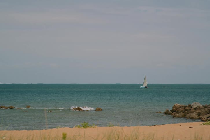 A lone sailboat glides across a calm blue sea near a sandy beach beneath a soft sky