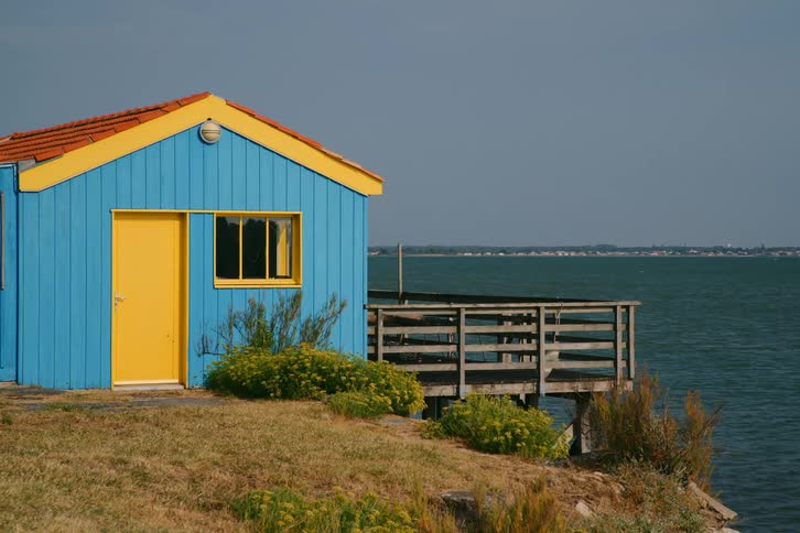 Blue house with a yellow door located by the seaside next to a wooden deck