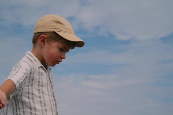 A young boy wearing a tan baseball cap and a plaid shirt walks outdoors while holding an adults hand against a backdrop of a blue sky with light clouds