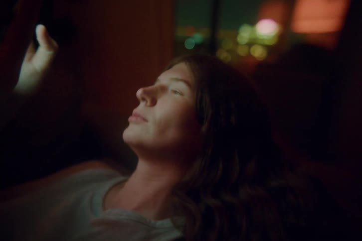 A woman with long brown hair lies in a dark room at night looking upward at her glowing smartphone screen while scrolling with her thumb