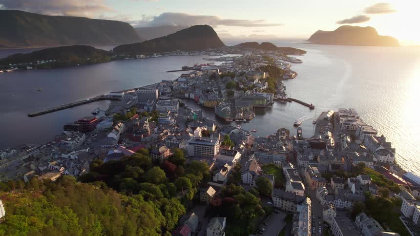 An aerial view captures a coastal city spread across narrow strips of land surrounded by deep blue water and mountain silhouettesthe golden light of a setting sun illuminates the dense clusters of buildings and harbors casting long shadows across th