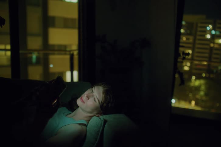 A woman reclines on a couch in a dark apartment at night scrolling through her glowing smartphone and behind her large windows reveal a blurred cityscape with illuminated office buildings and balconies under the night sky