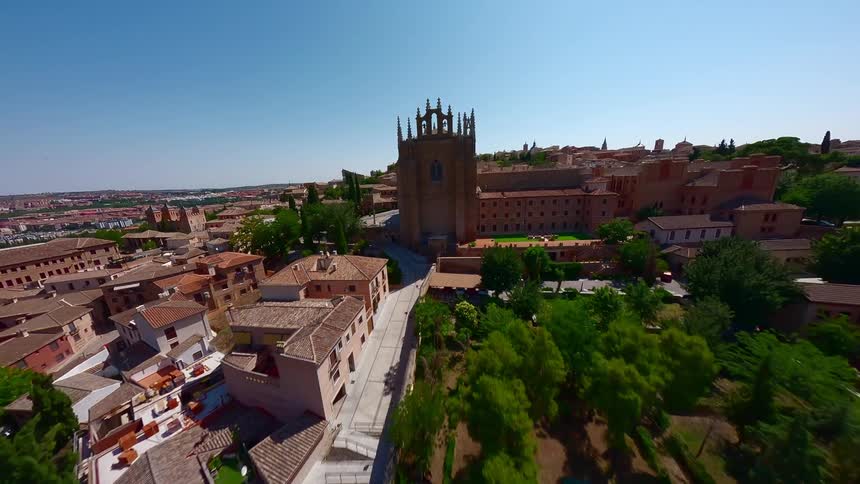 An aerial wide shot captures a dense historic city with brown tiled roofs and a large stone cathedral under a clear blue sky