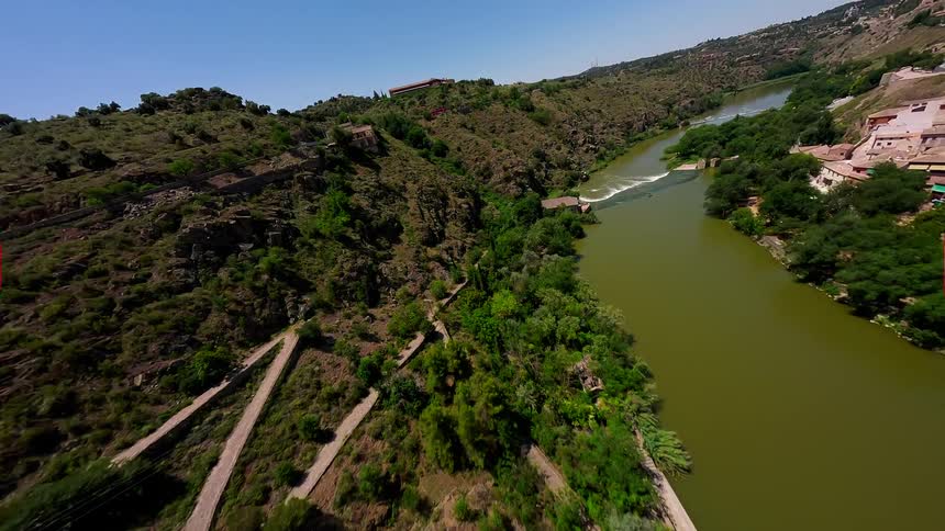 A drone flies over a rocky cliffside next to a green river revealing a winding stone path scattered mediterranean vegetation and a building perched on the hillside under a clear blue sky during a bright sunny day