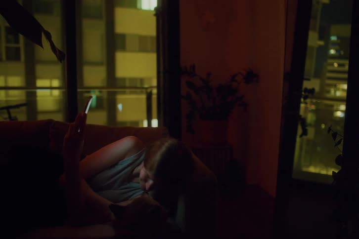 Woman lying on sofa at night looking at her cell phone and interacting with a dog