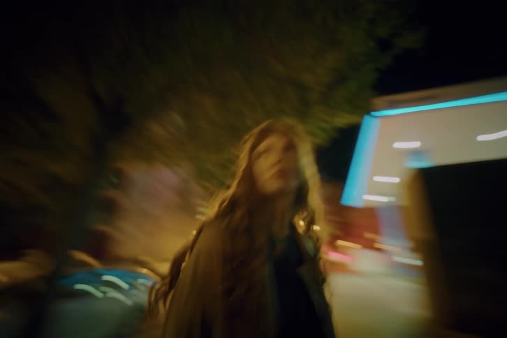 A young woman with long wavy hair spins around rapidly in the middle of a city street at night while blurred lights from buildings and traffic streak across the background