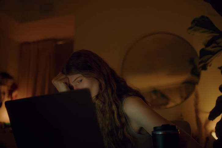 A young woman sits in a dimly lit room in front of a laptop while holding her hand to her forehead with her eyes closed in a gesture of fatigue or stress