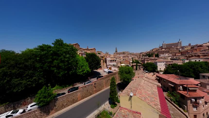 An aerial view flies over a green river towards a historic city on a hill before descending into a narrow alleyway between buildings