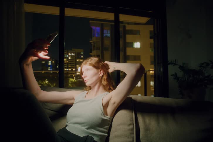 A woman sits on a sofa at night holding a smartphone and taking some selfies she adjusts her hair while looking at the screen with city buildings visible through the window behind her