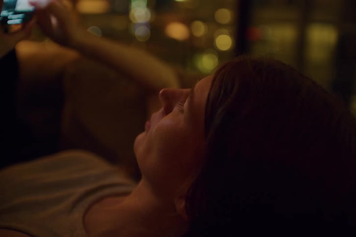 Person lying on a couch holds a lit smartphone in an indoor setting with blurred lights in the background