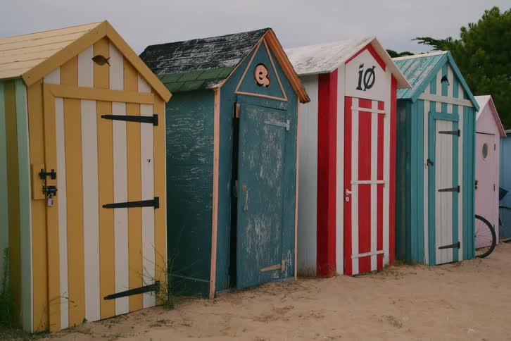 A row of colorful wooden beach huts painted with yellow blue and red vertical stripes stands on the sand near green trees