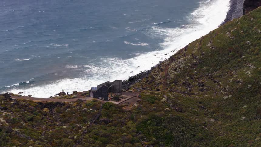 A small modern building with solar panels sits perched on a rocky green hillside overlooking a crashing coastlinea cable car descends toward the shoreline as white waves break against the dark volcanic rocks below
